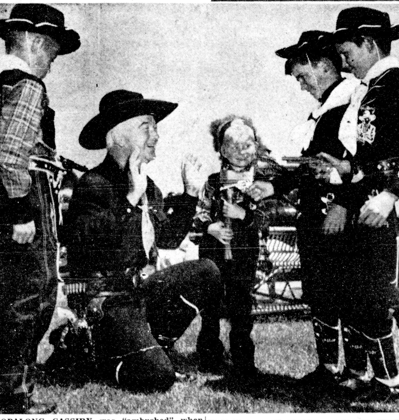 A newspaper photograph: Boyd in his black hat kneels on a lawn with his hands up, laughing, held up by four boys in cowboy outfits with toy guns and a small boy in a feathered headdress.