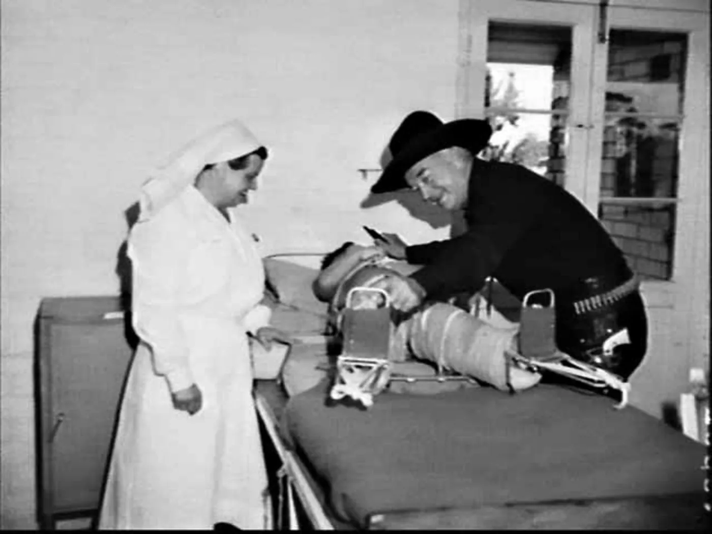In a ward by a window, Boyd in his black hat and gun belt bends over a small boy strapped into a spinal frame on a bed and shows him a toy gun; a nursing sister in a white veil smiles beside them.
