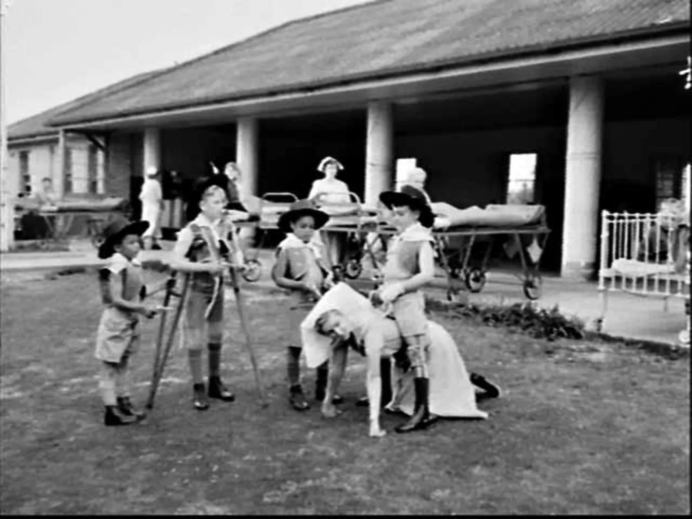 Five children in cowboy hats and neckerchiefs play at cowboys on a hospital lawn, two of them on crutches, with patients in beds along the verandah behind.