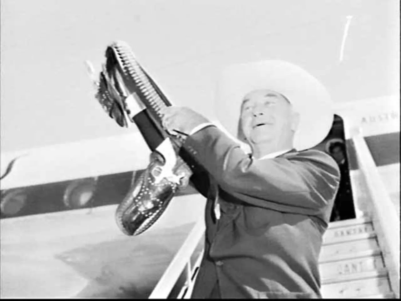 Boyd at the top of the aircraft steps in a white ten-gallon hat, shouting, holding up his gun belt and holstered pistol.