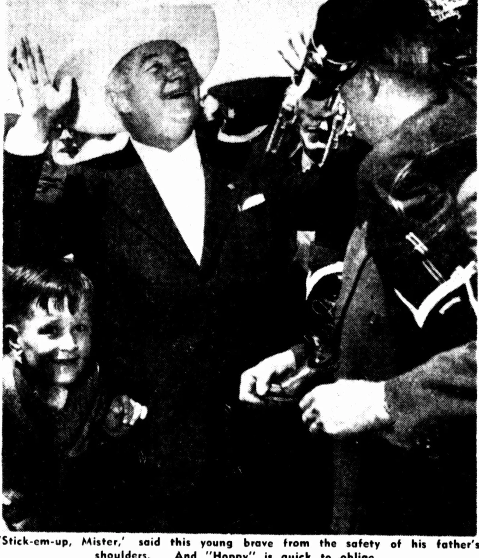 A newspaper photograph: Boyd in a white hat throws up both hands, laughing, held up by a small boy on his father’s shoulders. The printed caption: “‘Stick-em-up, Mister,’ said this young brave from the safety of his father’s shoulders. And ‘Hoppy’ is quick to oblige.”