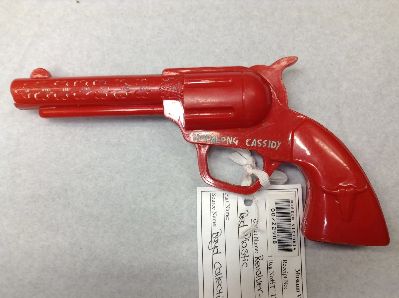 A red plastic toy revolver, “HOPALONG CASSIDY” moulded on its frame in silver and a steer’s head on the grip, with the museum’s tag tied to the trigger guard.